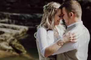 Ein Paar umarmt sich im Freien und lächelt sich herzlich an, während sie von ihrer Hochzeit träumen. Die Frau mit dem langen, welligen blonden Haar, das mit einem Blumenschmuck geschmückt ist, trägt ein weißes Kleid mit Spitzenärmeln. Ihr Partner trägt eine leichte Weste und ein Hemd. Sie stehen vor einer natürlichen, felsigen Kulisse.