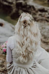 Eine Frau mit langen, welligen blonden Haaren sitzt im Freien, mit dem Rücken zur Kamera. Sie trägt eine leichte Weste und einen Faltenrock, perfekt für eine verträumte Hochzeitsplanungskulisse. Ihr Haar ist mit kleinen Blumen geschmückt, während sie einen Strauß rosa und weißer Blüten vor einem natürlichen, felsigen Hintergrund hält.