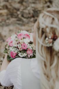 Eine Braut mit langen blonden Haaren, die mit Blumen gestylt sind, hält einen Strauß aus rosa und weißen Rosen und Grünpflanzen. Sie ist von hinten zu sehen und trägt ein helles Kleid, das vor einem unscharfen natürlichen Hintergrund perfekt die Essenz einer eleganten Hochzeit einfängt.