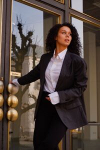 Eine Person mit lockigem Haar, für eine Hochzeit gekleidet in einem dunklen Blazer und einem weißen Hemd, öffnet eine Glastür mit einem „Ziehen“-Schild. Die Spiegelung von Bäumen und einem Gebäude tanzt auf dem Glas.