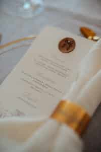Nahaufnahme einer eleganten Menükarte bei einer Hochzeit, die teilweise von einer weißen Serviette bedeckt ist, die von einem goldenen Ring gehalten wird. Das Menü enthält mehrere Gänge in anspruchsvoller Serifenschrift, während im Hintergrund ein goldenes Utensil glänzt und die Essenz sorgfältiger Hochzeitsplanung einfängt.