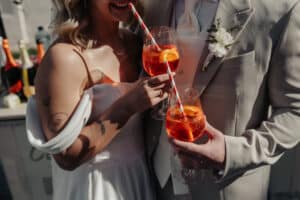 Braut und Bräutigam in eleganter Kleidung feiern ihre Hochzeit mit Orangencocktails, garniert mit Zitronenscheiben. Jedes Glas ist mit einem rot-weiß gestreiften Strohhalm ausgestattet. Sie stehen eng beieinander und zeigen einen freudigen und intimen Moment der Zweisamkeit.
