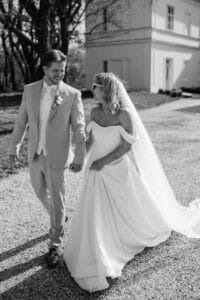 Ein Paar geht Hand in Hand auf einem Kiesweg vor einem weißen Gebäude und fängt die Essenz ihrer Hochzeit ein. Der Bräutigam trägt einen hellen Anzug und die Braut ein schulterfreies Kleid mit langem Schleier und Sonnenbrille. Beide lächeln, während sie gehen, umgeben von Bäumen. Schwarzweißfoto.