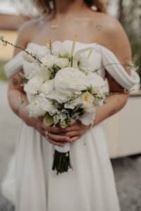 Eine Braut in einem schulterfreien weißen Kleid, perfekt für eine stilvolle Hochzeit, hält einen Strauß aus weißen Rosen, Callas und Grünpflanzen. Sie steht im Freien mit tätowierten Armen, auf deren Schulter ein kleines Herz zu sehen ist. Der Hintergrund ist leicht verschwommen.