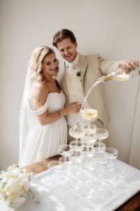 Braut und Bräutigam in Abendgarderobe gießen bei ihrer Hochzeit fröhlich Champagner in eine Pyramide aus Gläsern. Ein Strauß weißer Blumen steht daneben und unterstreicht die elegante Kulisse.
