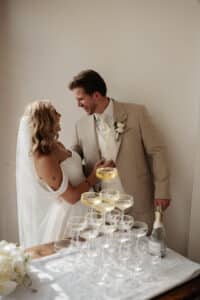 Braut und Bräutigam stehen neben einer Pyramide aus Champagnergläsern und verkörpern damit die Freude ihrer Hochzeit. Die Braut in einem schulterfreien weißen Kleid und Schleier lächelt den Bräutigam im beigen Anzug an, der eine Flasche hält. Sonnenlicht fällt durch und schafft eine warme Atmosphäre, die perfekt zum Heiraten ist.