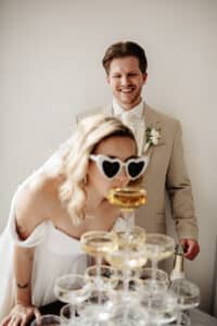 Ein Paar bei seiner Hochzeit. Die Frau trägt ein weißes Kleid und eine herzförmige Sonnenbrille und nippt an einem Champagnerturm. Der Mann im beigen Anzug steht lächelnd im Hintergrund neben einer Champagnerflasche und schafft damit die perfekte Kulisse für ihre Hochzeitsfeier.