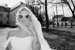 Eine Braut in einem schulterfreien Kleid hält einen transparenten, mit Perlen verzierten Schleier vor dem Gesicht und trägt eine herzförmige Sonnenbrille. Tattoos zieren ihren Arm, während sie draußen neben einem großen Gebäude und Bäumen steht. Das Schwarz-Weiß-Bild fängt die Essenz der Eleganz und Kunstfertigkeit einer Hochzeit ein.
