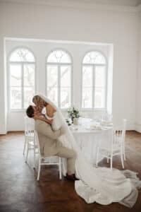 Braut und Bräutigam küssen sich vor einem runden Tisch mit elegantem Geschirr vor großen Bogenfenstern. Der lange Schleier und das Kleid der Braut liegen auf dem Holzboden, was die Essenz der Hochzeitsplanung widerspiegelt. Der Raum ist hell und wunderschön beleuchtet.