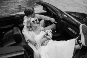 Ein Paar sitzt fröhlich in einem Cabrio. Die Frau trägt ein Brautkleid und eine herzförmige Sonnenbrille und hält einen Blumenstrauß in der Hand. Der Mann trägt einen Anzug und lächelt verspielt. Ganz im Geiste einer Hochzeit verkörpern sie einen unbeschwerten und feierlichen Moment an ihrem besonderen Tag.