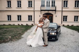 Braut und Bräutigam umarmen sich vor einem eleganten Gebäude und lächeln an ihrem Hochzeitstag. Sie trägt ein weißes Kleid und hält einen Blumenstrauß in der Hand. Er trägt einen hellen Anzug. Hinter ihnen auf der Kiesauffahrt parkt ein schwarzer Oldtimer, Teil ihrer sorgfältigen Hochzeitsplanung.