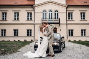 Ein Paar küsst sich vor einem schwarzen Oldtimer auf einem Schotterweg. Der Bräutigam im beigen Anzug und die Braut im weißen Kleid mit langer Schleppe genießen den Moment, eingerahmt von einem großen cremefarbenen Gebäude mit rotem Dach – eine idyllische Szene direkt aus ihrer Hochzeitsplanung.