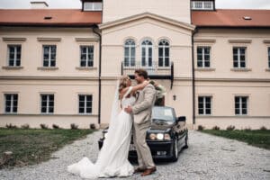 Braut und Bräutigam umarmen sich vor einem schwarzen Auto auf einer Kiesauffahrt und halten damit einen perfekten Hochzeitsmoment fest. Hinter ihnen steht ein elegantes, helles Gebäude mit rotem Dach. Die Braut trägt ein langes weißes Kleid, während der Bräutigam in seinem hellbraunen Anzug elegant aussieht und die Essenz perfekter Hochzeitsplanung verkörpert.