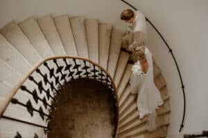 Braut und Bräutigam steigen eine Wendeltreppe hinab und verkörpern damit die Eleganz einer sorgfältig orchestrierten Hochzeitsplanung. Die Braut in ihrem fließenden weißen Kleid mit dem Blumenstrauß in der Hand ergänzt den beigen Anzug des Bräutigams. Sie gleiten die Stufen hinunter, die sich anmutig unter einem schmiedeeisernen Geländer winden.
