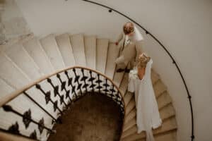 Ein Paar geht anmutig eine Wendeltreppe aus Stein hinunter. Die Person links trägt einen beigen Anzug und die Person rechts, strahlend in ihrem weißen Hochzeitskleid, hält Blumen in der Hand. Die Szene ihrer Hochzeit entfaltet sich wunderschön vor dem schwarzen schmiedeeisernen Geländer.