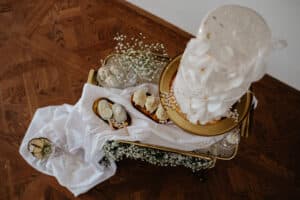 Ein dekoratives Arrangement für eine Hochzeit mit einer hohen, eleganten weißen Torte, die mit Blumenmustern geschmückt ist. Sie steht elegant auf einem goldenen Tablett mit dazugehörigem Dekor wie kleinen Desserts, Blumen und einem weißen Tischtuch, alles auf einem strukturierten Holzboden – ideal für Ihre Hochzeitsplanung.