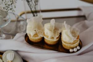 Drei elegante Gebäckstücke, belegt mit zarter Sahne und dekorativen Oblatenstücken, liegen auf einem goldenen Tablett. Der Hintergrund ist perfekt für Hochzeitsfeierlichkeiten und umfasst ein weiches weißes Tuch und Blumenakzente in einem minimalistischen und raffinierten Arrangement, das die Essenz raffinierter Hochzeitsplanung verkörpert.