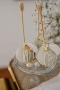 Zwei elegante weiße Cake Pops am Stiel, verziert mit Blattgold und weißen Perlen, werden auf einem Glasständer präsentiert. Zarte weiße Blumen verschwimmen im Hintergrund und verleihen dieser exquisiten Hochzeitspräsentation einen Hauch von Anmut.