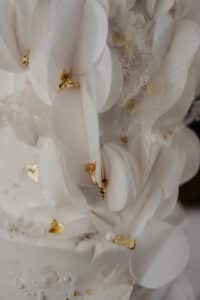 Nahaufnahme einer eleganten weißen Hochzeitstorte, die mit zarten weißen Blütenblättern und Blattgoldakzenten verziert ist. Perfekt für diejenigen, die gerade mit der Hochzeitsplanung beschäftigt sind. Die Torte weist komplizierte Texturen auf und sieht luxuriös und stilvoll aus, was sie zur idealen Wahl für eine anspruchsvolle Hochzeit macht.