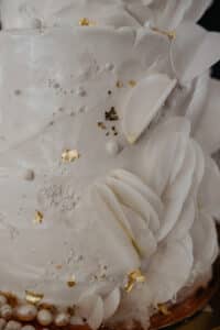 Nahaufnahme einer eleganten weißen Hochzeitstorte, die mit zarten weißen Blütenblättern und Blattgoldakzenten geschmückt ist. Die Zuckergusstextur ist glatt mit subtilen dekorativen Mustern, was ein anspruchsvolles und luxuriöses Erscheinungsbild erzeugt, perfekt für jede Hochzeitsfeier.