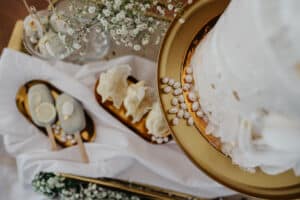 Eine weiße Schichttorte mit Blumendekorationen auf einem goldenen Ständer, perfekt für jede Hochzeit. Dazu gibt es elegant Cake Pops und Cracker auf einem Tablett. Daneben sind Schleierkrautblüten liebevoll arrangiert, alles auf einem makellos weißen Tuch platziert, was eine ideale Hochzeitsszene ergibt.