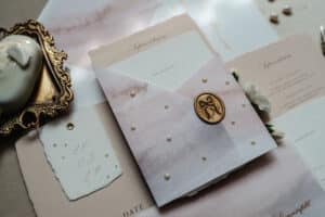 Eine Sammlung eleganter Hochzeitspapeterie mit Aquarellmotiven und Perlenakzenten, perfekt für jede Hochzeit. Es gibt einen Umschlag mit einem monogrammierten Wachssiegel, umgeben von floralen Elementen und einem dekorativen Goldrahmen mit Blumen. Ideal für alle, die sich intensiv mit der Hochzeitsplanung beschäftigen.