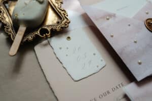 Eine flache Lage eleganter Briefpapiere für die Hochzeitsplanung, darunter eine „Save the Date“-Karte mit dem Datum „26 03 24“, verziert mit kleinen Perlen. Die Karte liegt auf einer strukturierten Oberfläche neben einem verzierten Rahmen und einem cremigen Eis am Stiel, perfekt für diejenigen, die heiraten möchten.