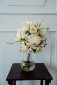 Ein Strauß weißer Blumen, darunter Rosen und Lilien, arrangiert in einer klaren geriffelten Glasvase, perfekt für eine Hochzeit. Die Vase steht elegant auf einem dunklen Holztisch vor einer hell getäfelten Wand.