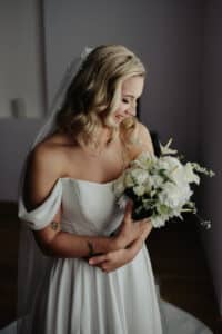 Eine Braut in einem schulterfreien Brautkleid hält einen Strauß weißer Blumen und verkörpert damit die Eleganz einer sorgfältig geplanten Hochzeit. Lächelnd und nach unten blickend fällt ihr welliges blondes Haar über ihre Schultern, und ein weicher Schleier mit kleinen Perlenakzenten fällt anmutig hinter ihr herab.