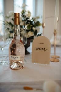 Eine Flasche Roséwein mit der Aufschrift „Studio“ auf einer weißen Tischdecke, umgeben von elegantem Glasgeschirr, mit einer beigen Karte mit der Aufschrift „Tisch vier“ und einem Blumengesteck im Hintergrund verleihen Ihrer Hochzeitsfeier den perfekten Touch.