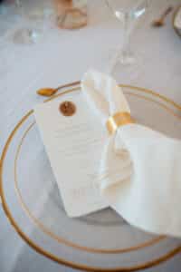 Die elegante Hochzeitstafel besteht aus einem transparenten Glasteller mit Goldrand. Auf dem Teller liegen ein gefaltetes Menü mit Wachssiegel und eine elfenbeinfarbene Serviette, die in ein goldenes Papierband eingewickelt ist. Daneben liegt eine Gabel mit goldenem Griff auf der weißen Tischdecke und spiegelt die Essenz anspruchsvoller Hochzeitsplanung wider.