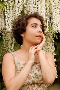 Eine Person mit kurzen, lockigen Haaren und einem geblümten Kleid steht vor herabhängenden weißen Blumen und berührt sanft ihr Gesicht mit einer Hand. Sie wirkt nachdenklich und denkt über Hochzeitspläne nach, während sie nach oben rechts blickt.