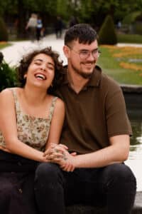 Eine Frau und ein Mann sitzen eng beieinander an einem Teich, lächeln und halten Händchen, während sie von ihrer bevorstehenden Hochzeit träumen. Die Frau hat lockiges Haar und trägt ein geblümtes Oberteil, während der Mann eine Brille und ein braunes Hemd trägt. Ein Pfad und viel Grün bieten die perfekte Kulisse für ihre Liebesgeschichte.