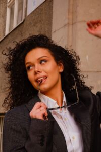 Eine Person mit lockigem Haar, einem dunklen Blazer und einem weißen Hemd, einer Sonnenbrille in der Hand und einem verspielten Gesichtsausdruck, scheint von ihrer bevorstehenden Hochzeit zu träumen. Sie scheint draußen an einem Gebäude zu stehen.