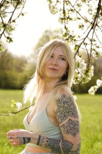 Eine Frau mit Tattoos steht auf einem Feld, umgeben von blühenden Zweigen, und träumt von ihrer bevorstehenden Hochzeit. Ihr langes blondes Haar fällt über ein helles Oberteil, während sie unter einem klaren Himmel in die Ferne blickt und in Gedanken über Liebe und Hochzeitsplanung versunken ist.