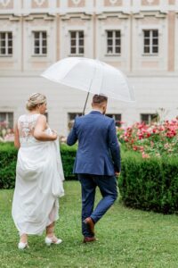 In einer ruhigen Gartenlandschaft gehen Braut und Bräutigam über üppiges Gras. Der Bräutigam hält einen großen weißen Regenschirm, der das weiße, mit Spitzendetails verzierte Kleid der Braut ergänzt. In einem blauen Anzug sticht er zwischen dem leuchtenden Grün und den Blumen hervor – ein bildschöner Moment ihrer Hochzeit.