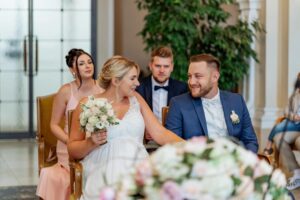 Eine Braut in einem weißen Kleid hält einen Blumenstrauß und lächelt den Bräutigam im blauen Anzug während ihrer Hochzeit an. Zwei Trauzeugen sitzen hinter ihnen, einer in einem rosa Kleid und der andere in einem dunklen Anzug, während üppiges Grün die Kulisse wunderschön schmückt.