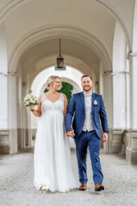 Ein Paar geht freudig Hand in Hand unter einem eleganten Torbogen hindurch und verkörpert damit die Romantik ihrer Hochzeit. Die Braut trägt ein weißes Kleid und hält einen Blumenstrauß, während der Bräutigam einen blauen Anzug mit weißer Weste trägt. Die helle Umgebung unterstreicht diese wunderschön geplante Feier der Liebe.