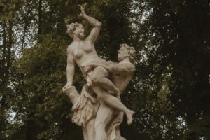 Eine Marmorstatue, die zwei ineinander verschlungene Figuren in einer dynamischen Pose zeigt, die an einen eleganten Hochzeitstanz erinnert, vor einem Hintergrund aus üppigen grünen Bäumen. Die Figur auf der linken Seite scheint eine Verteidigungshaltung einzunehmen, während die andere Figur nach ihnen greift.