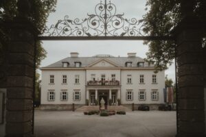 Hinter den verzierten schwarzen schmiedeeisernen Toren erwartet Sie ein großes, elegantes weißes Herrenhaus, das perfekt für eine Traumhochzeit ist. Die symmetrische Fassade verfügt über mehrere Fenster und einen Balkon mit Blumentöpfen. Davor parken zwei Autos, ideal für diejenigen, die ihren besonderen Tag planen, während Bäume die Szene wunderschön umrahmen.