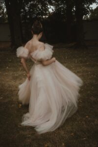 Eine Person trägt ein fließendes, elegantes, blassrosa Kleid mit Rüschenärmeln und läuft während einer Hochzeit über Gras. Die Person hat ihr Haar zu einem Knoten hochgesteckt und ist von Bäumen umgeben, was eine heitere, ätherische Atmosphäre schafft, die perfekt für eine Hochzeit ist.