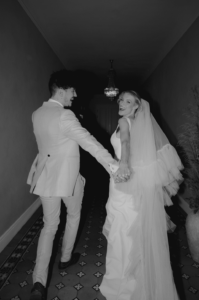 Braut und Bräutigam, vertieft in ihre Hochzeitsplanung, halten freudig Händchen, während sie einen gemusterten Flur entlanggehen. Das fließende Kleid und der lange Schleier der Braut ergänzen den hellen Anzug des Bräutigams, während sie lächelnd unter einem Kronleuchter zurückblicken.