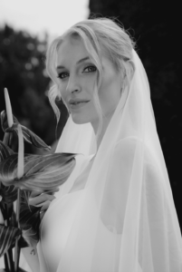 Eine Frau mit hellem Haar in einem eleganten Brautkleid und Schleier steht draußen und verkörpert einen Moment der Hochzeitsmagie. Sie hält einen Strauß mit großen Blättern, lächelt sanft, mit einem unscharfen natürlichen Hintergrund hinter ihr. Schwarzweißbild.