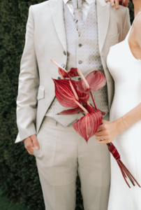 Ein Paar in formeller Kleidung steht zusammen bei seiner Hochzeit, der Mann trägt einen hellgrauen Anzug und die Frau ein weißes Kleid. Sie hält einen Strauß roter Anthurienblüten. Sie stehen draußen mit grünem Laub im Hintergrund und fangen die Essenz der Liebe an ihrem besonderen Tag ein.