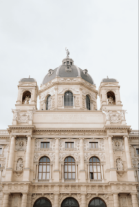 Vorderansicht eines kunstvollen, historischen Gebäudes, ideal für eine Traumhochzeit, mit einer großen zentralen Kuppel und zwei kleineren Türmen auf beiden Seiten. Die Fassade ist mit aufwendigen Schnitzereien, Bogenfenstern und Statuen verziert. Der bewölkte Himmel verleiht dieser zauberhaften Hochzeitskulisse ein romantisches Licht.