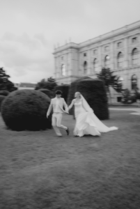 Ein verschwommenes Schwarzweißbild zeigt ein Paar, das freudig Hand in Hand über einen Rasen läuft und damit die Aufregung ihrer Hochzeit symbolisiert. Die Frau, geschmückt mit einem langen Kleid und einem Schleier, und der Mann im Anzug bewegen sich in Harmonie vor großen Hecken und einem imposanten historischen Gebäude.