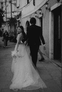 Ein Paar geht Hand in Hand eine malerische Straße entlang, eingehüllt in die Glückseligkeit seiner Hochzeit. Die Frau, in einem fließenden Brautkleid, blickt über ihre Schulter zurück und hält einen Blumenstrauß in der Hand, während der Mann, für ihren besonderen Tag herausgeputzt, den Weg anführt. Straßenlaternen und Fußgänger bilden eine bezaubernde Kulisse.