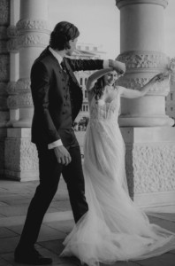 Ein Schwarzweißfoto fängt die Freude einer Hochzeit ein, während ein Paar im Freien tanzt. Die Frau strahlt in einem langen Spitzenhochzeitskleid, während ihr Bräutigam im Anzug sie anmutig in der Nähe verzierter Steinsäulen wirbelt.