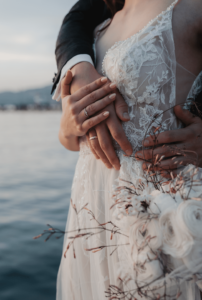 Ein Paar umarmt sich an einem ruhigen Ufer und feiert seine Hochzeit. Die Braut trägt ein Spitzenhochzeitskleid und hält einen Strauß weißer Rosen in der Hand. Die Arme des Bräutigams schlingen sich um sie und präsentieren ihre Eheringe vor dem Hintergrund des ruhigen Wassers unter einem sanften Himmel.