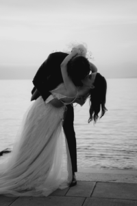 Ein Paar im Hochzeitskleid küsst sich bei Sonnenuntergang am Meer. Der Bräutigam taucht die Braut ins Wasser, deren langes Kleid elegant herabfällt. Hochzeitsglück liegt in der Luft, ein ruhiges Meer und ein sanft beleuchteter Horizont bilden die Kulisse und fangen einen romantischen und zeitlosen Moment ein, der sich perfekt als Inspiration für jede Hochzeitsplanung eignet.